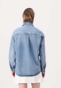 Giacca in denim di un blu chiaro con maniche lunghe, dotata di colletto classico, piega sul retro e polsini con bottone. L'orlo arrotondato aggiunge un tocco di dettaglio.
