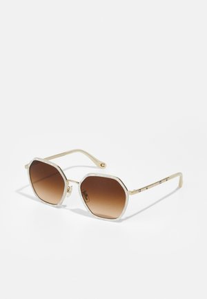 Gafas de sol octagonales con montura crema y lentes degradados en marrón. Presenta acentos metálicos en las patas y un diseño texturizado en la montura.