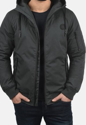 Man draagt een donkergrijze capuchonjas over een zwart shirt, met handen in de voorzakken, staand tegen een effen achtergrond.