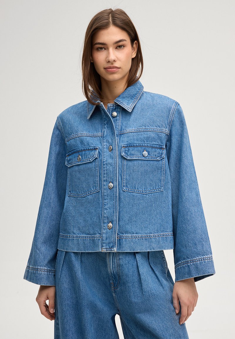 Junge Frau trägt eine hellblaue Jeansjacke mit geknöpften Brusttaschen und passende, hoch taillierte Jeanshosen.