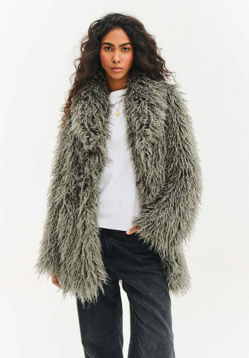 Kobieta o długich kręconych włosach nosi dużą szarą, puszystą futrzaną kurtkę z futra syntetycznego, białą koszulę, czarne spodnie i nawarstwione złote naszyjniki, stojąc na białym tle.