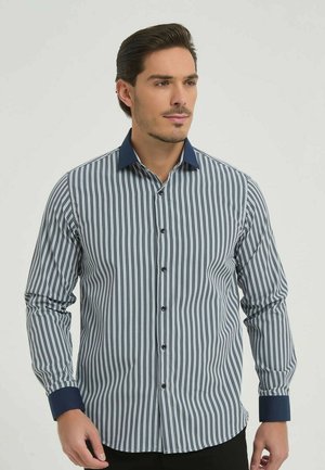 Homme portant une chemise à manches longues à rayures verticales avec col et poignets bleu foncé, se tenant devant un fond uni.