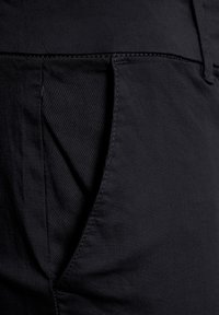 Gros plan sur un pantalon en tissu sombre montrant une poche latérale et des coutures détaillées.