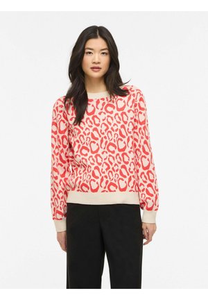 Vrouw met lang donker haar, die een beige en rode trui met luipaardprint en zwarte broek draagt, staand tegen een witte achtergrond.