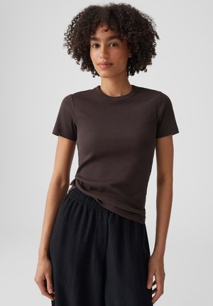 Junge Frau mit lockigem Haar, die ein eng anliegendes dunkelbraunes Kurzarmshirt und lockere schwarze Hose vor einem schlichten Hintergrund trägt.