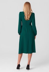 Zielona sukienka do kolan z długimi, bufiastymi rękawami i dopasowanym pasem. Gładka tekstura, jednolity kolor i subtelne detale szwów z tyłu.