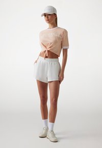 Persiko kortärmad knyt-t-shirt med vita detaljer och logga, kombinerad med vita shorts med elastisk midja och vita sneakers, mot en ljus bakgrund.