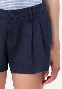 Ναυτικό μπλε σορτς με πλισέ μπροστά, δύο πλάγιες τσέπες και κλείσιμο με κουμπί. Φτιαγμένο από ελαφρύ ύφασμα, με λεία υφή.