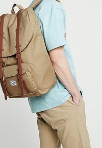 Beżowy plecak z płótna z brązowymi skórzanymi paskami, z zamknięciem na klapkę i przednią kieszenią. Noszony na ramieniu, w połączeniu z jasnoniebieską koszulą.