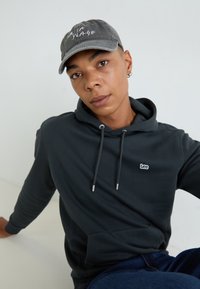 Donkergrijze hoodie met trekkoorden en een voorzak, uitgerust met een klein wit "Lee" logo. Gecombineerd met een grijze pet met de tekst "La Plage".