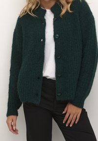 Cardigan verde scuro realizzato in tessuto a maglia testurizzato, con scollatura rotonda e bottoni neri sulla parte anteriore. Indossato su una camicia bianca.