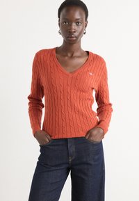 V-neck, cable-knit sweater in burnt orange wool blend, featuring long sleeves and a subtle logo on the chest. Paired with dark jeans.