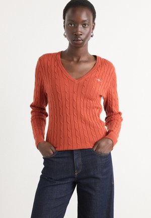 V-hals, kabelstrikket genser i brent oransje ullblanding, med lange ermer og en subtil logo på brystet. Kombinert med mørke jeans.