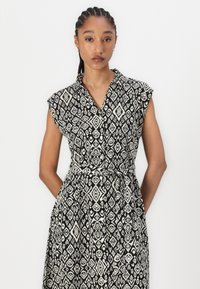 Rochie cămașă cu un model alb-negru, cu mâneci scurte, cu un design cu butoni în față și o curea asortată la talie. Motive în formă de diamant pe întreaga suprafață.