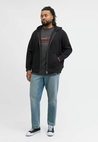 Felpa nera con zip sopra una t-shirt grafica grigia, abbinata a jeans blu chiaro e sneaker nere, con suole bianche e lacci neri.