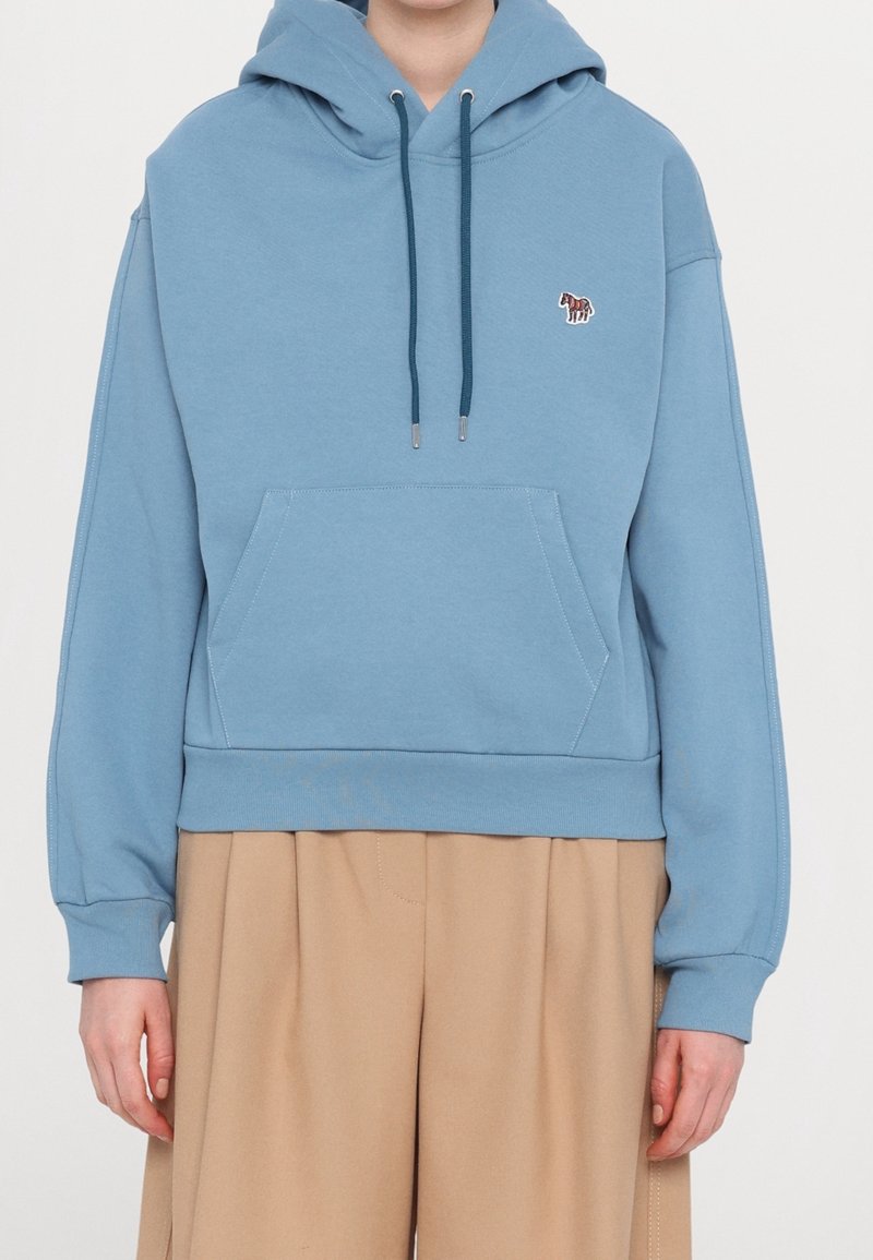 Personne portant un sweat à capuche bleu clair avec une poche sur le devant et un petit patch zèbre sur la poitrine, associé à un pantalon beige à jambes larges.