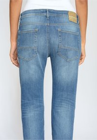 Persona que lleva jeans de mezclilla azul claro con bolsillos traseros y una etiqueta de marca de cuero beige en la cintura, vista desde atrás.