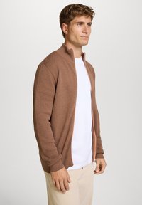 Brun tekstureret zip-jakke med høj krave, kontrasteret af en orange lynlås, båret over en hvid skjorte og lyse beige bukser.