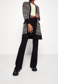 Černobílý vzorovaný kardigan s kapsami, nošený přes zelený crop top. Kombinován s černými širokými kalhotami s žebrovaným vzorem a robustními černými botami.