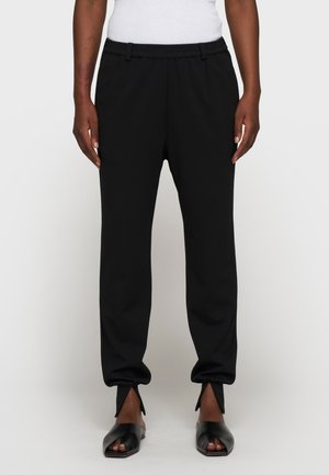 Persona vistiendo pantalones negros ajustados con aberturas delanteras en los tobillos y zapatos negros de tacón abiertos en los dedos, con una blusa blanca sin mangas visible.