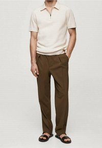 Homme portant un polo en tricot beige à manches courtes, un pantalon plissé marron et des sandales noires, se tenant avec une main dans sa poche devant un fond uni.