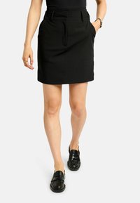 Fekete pamut szoknya magas derékkal, pliszírozott elejjel és oldalzsebekkel. Fekete mokaszinokkal viselve, térd alatti hosszúságú kialakítással.