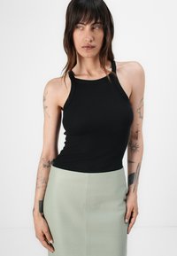 Svart ribbet tank topp med dyp utringning og tynne stropper, kombinert med en lys grønn tettsittende skjørt. Tatoveringer synlige på armene.
