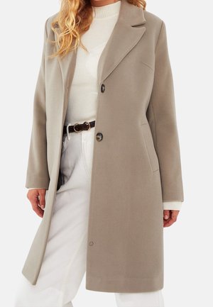 Manteau en laine beige pour femme avec deux boutons, porté sur un pull à col roulé côtelé blanc et un pantalon blanc avec une ceinture marron.