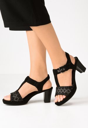 Czarne zamszowe sandały na obcasie z odkrytymi palcami, regulowanym paskiem wokół kostki oraz ozdobionym przednim panelem z metalicznymi akcentami.