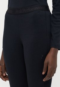 Leggings de sport noirs avec une large taille arborant la marque "PeakPerformance". Texture lisse avec un léger éclat. Ajustement mi-haut.