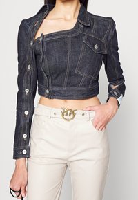 Denimjacka med avskuren längd i mörk tvätt och asymmetrisk design, framfickor och kontrasterande sömmar; kombinerad med beige byxor och en utsmyckad bälte.