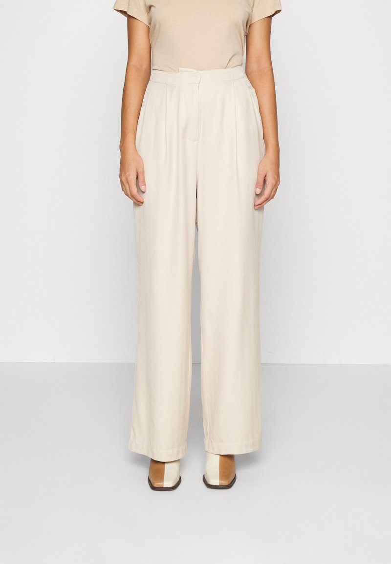 Pantalon beige taille haute à jambes larges avec des plis et une texture lisse, associé à des chaussures beige clair avec un design géométrique.