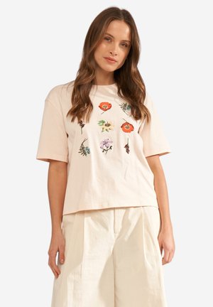 Mujer vistiendo una camiseta beige claro de gran tamaño con flores bordadas de colores, combinada con pantalones sueltos de color crema.