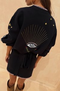 Čierny pletený odev s nadrozmernými rukávmi, ktorý obsahuje vyšívaný dizajn oka a zlaté hviezdne akcenty. S previazaným pásom a kontrastnými detailmi na spodnom okraji.