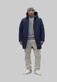 Chaqueta de plumas azul marino con bolsillos con cremallera, suéter de cuello alto gris, pantalones beiges y zapatillas blancas. Un gorro gris completa el look.