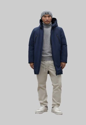 Chaqueta de plumas azul marino con bolsillos con cremallera, suéter de cuello alto gris, pantalones beiges y zapatillas blancas. Un gorro gris completa el look.