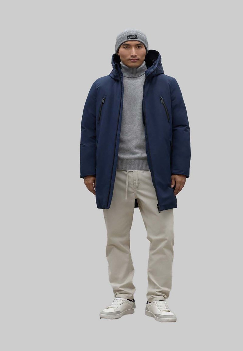 Chaqueta de plumas azul marino con bolsillos con cremallera, suéter de cuello alto gris, pantalones beiges y zapatillas blancas. Un gorro gris completa el look.