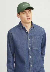Denim skjorte med knapper i mørkeblå med en frontlomme og hvide knapper, parret med en grøn kasket med hvid skrift.