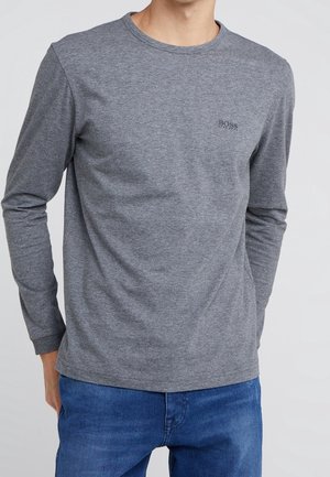 Muž v jednoduchém šedém tričku s dlouhým rukávem s malým logem "BOSS" na hrudi a v modrých džínách na světlém pozadí.