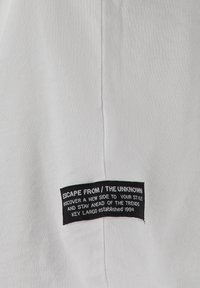 Etiqueta de tela negra en prenda blanca con texto blanco: "ESCAPE DE / LO DESCONOCIDO DESCUBRE UN NUEVO LADO DE TU ESTILO Y MANTENTE A LA VANGUARDIA DE LAS TENDENCIAS KEY LARGO establecido en 1994".