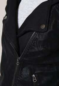 Giubbotto in pelle nera con chiusura a zip, grande colletto e dettagli in tessuto a contrasto. Finitura testurizzata con dettagli in metallo color argento.