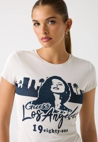 Biele tričko s krátkym rukávom, ktoré zobrazuje modrý grafický dizajn siluety ženy a text "Guess Los Angeles 1981", vyrobené z jemnej bavlny.
