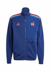 Blaue Zip-Jacke aus weichem Material mit Stehkragen. Mit roten und weißen Streifen auf den Schultern und einem Hahn-Logo auf der Brust.