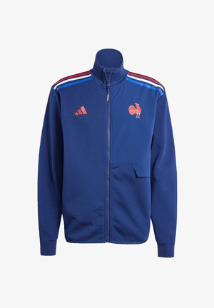 Giacca blu con zip realizzata in materiale morbido e colletto rialzato. Presenta righe rosse e bianche sulle spalle e un logo di gallo sul petto.