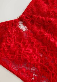 Rote Spitzenstoff mit filigranen Blumenmustern, einer gewellten Kante und einem goldenen, kreisförmigen Ringdetail am Halsausschnitt.