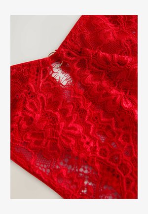 Červená krajková látka s jemnými květinovými vzory, zakončená vroubkovaným okrajem a zlatým kruhovým prstencem u výstřihu.
