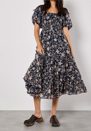 Femme portant une robe midi bleu marine à manches bouffantes avec un imprimé floral beige, associée à des sabots marron foncé à talons en bois et clous.