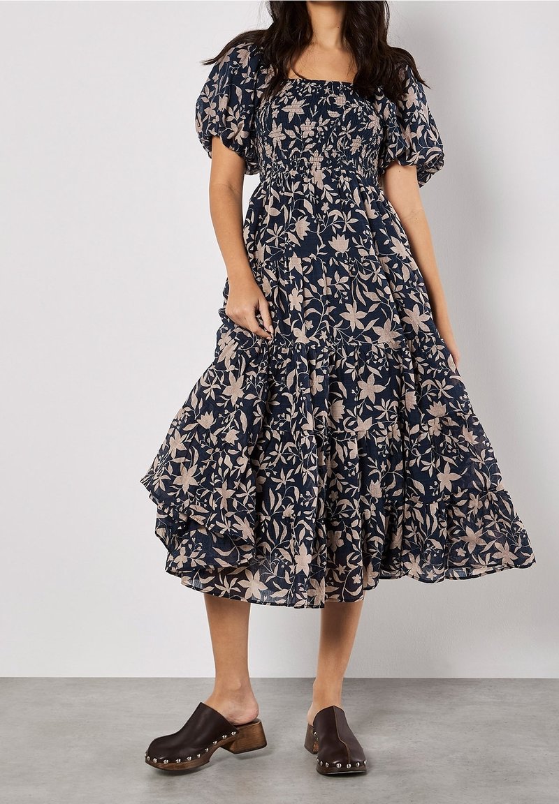 Femme portant une robe midi bleu marine à manches bouffantes avec un imprimé floral beige, associée à des sabots marron foncé à talons en bois et clous.
