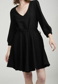 Svart A-linje klänning med djup V-hals, långa puffärmar och justerbart midjeband. Tillverkad av mjukt tyg, har den två sidofickor.