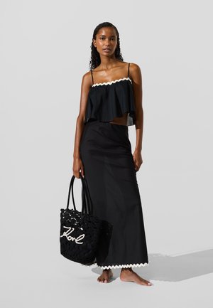 Crni kratki top s bijelim valovitim rubom, uparen s dugom crnom suknjom. Drži crnu ručno rađenu torbu s bijelim natpisom. Bosa.